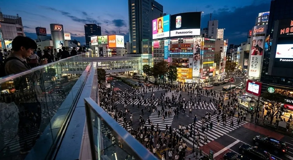 A high-angle twilight view of Shibuya Crossing from an elevated glass observation deck, showing thousands of people crossing the illuminated streets amidst vibrant neon skyscrapers.
