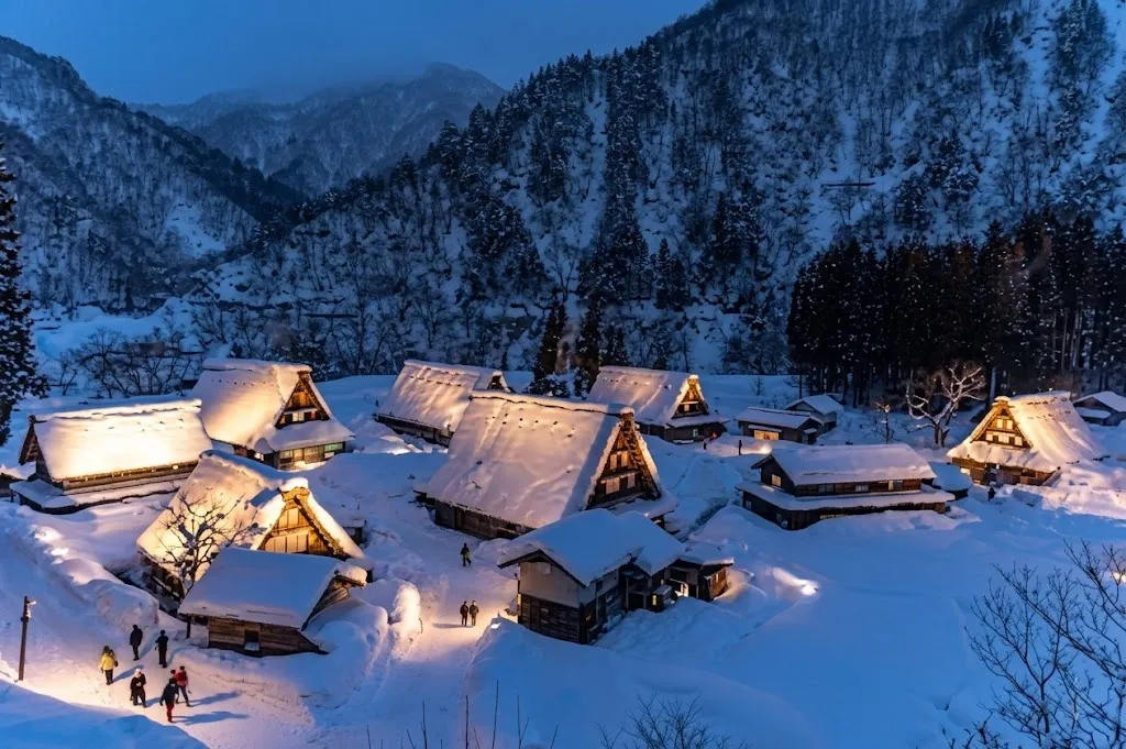 Traditional Japanese thatched-roof farmhouses glow warmly while nestled under deep, heavy blankets of white snow in a peaceful UNESCO World Heritage mountain village at the onset of twilight.